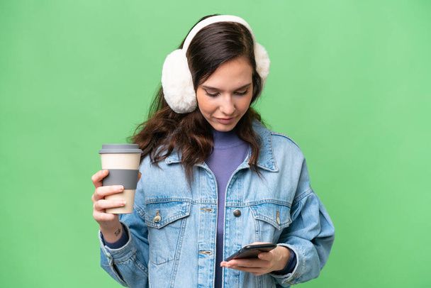 Young caucasian woman wearing winter muffs over isolated background holding coffee to take away and a mobile - 写真・画像