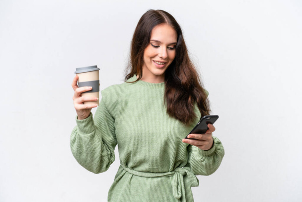 Young caucasian woman isolated on white background holding coffee to take away and a mobile - Photo, Image
