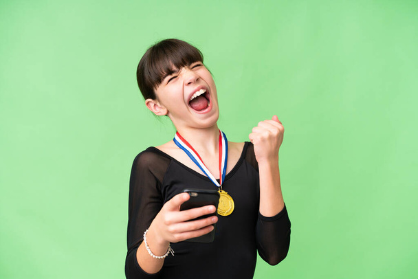 Pequeña chica caucásica con medallas sobre fondo aislado con teléfono en posición de victoria - Foto, Imagen