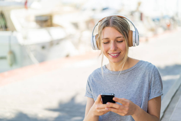 Fiatal szőke nő a szabadban zenét hallgat, és keresi a mobil - Fotó, kép