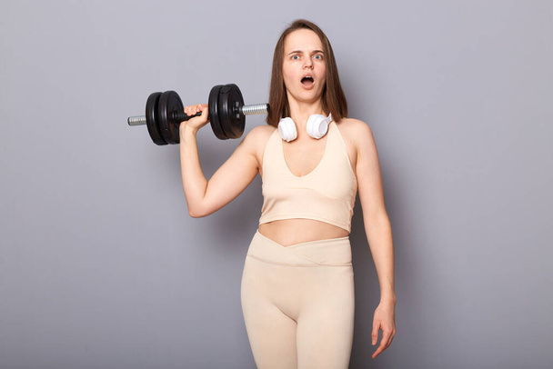 Shocked surprised woman wearing top holding red barbells in hands isolated on gray background looking ta camera with open mouth raised arm with dumbbell. - Foto, Imagen