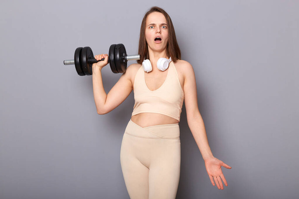 Sporty woman wearing beige top and leggins holding barbel pumps biceps and triceps leads healthy lifestyle posing isolated over gray background looking at camera with open mouth. - Photo, Image