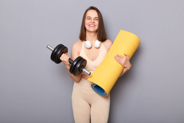 Cheerful attractive athletic woman with yoga mat and dumbbell posing isolated over gray background looking at camera leads healthy lifestyle - Fotó, kép