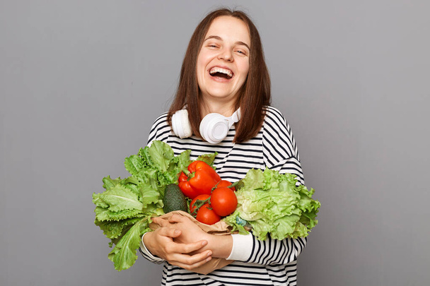 Green diet, healthy living. Laughing woman holding vegetables isolated over gray background. Vegetarian lifestyle, health choice. Fresh vegetables, nutritious boost. - Фото, изображение