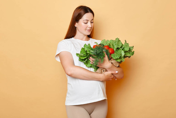 Future mother's nutrition. Green market for health. Adult brunette attractive pregnant woman wearing white shirt holding fresh vegetables posing isolated over beige background - Zdjęcie, obraz