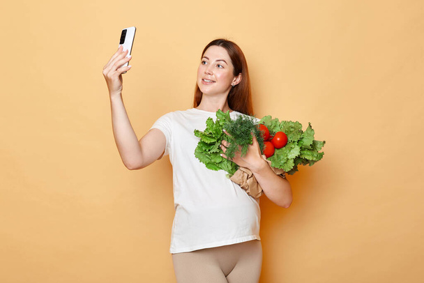 Beautiful attractive pregnant woman holding fresh vegetables posing isolated over beige background making selfie on cell phone holding mobile phone. - Foto, Bild