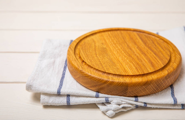 Empty wooden round pizza board on kitchen white wooden table. Cutting board with red checkered kitchen towel. Wooden pizza plate, copy space. Kitchenware. Recipe. Place for text. Flat lay. - Photo, Image