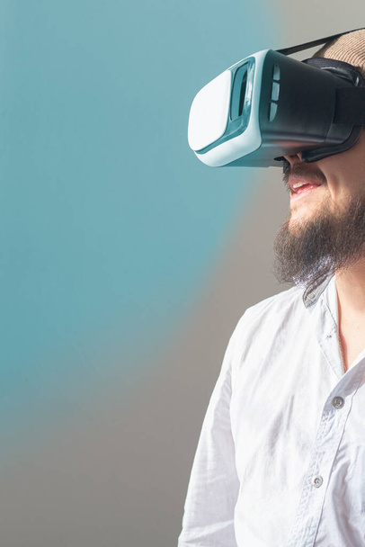 Young bearded man smiling using VR glasses to access the internet. Virtual reality glasses projecting cyan light. - Foto, Imagen