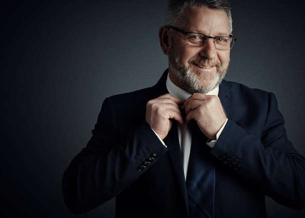 I wear my tie with confidence. Studio portrait of a handsome mature businessman fastening his tie while standing against a dark background - Φωτογραφία, εικόνα