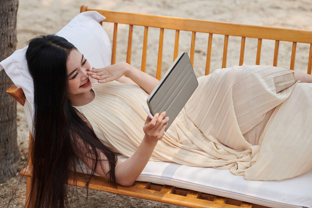 Молодая женщина отдыхает на скамейке на песчаном пляже и смотрит новый эпизод ситкома на планшетном компьютере - Фото, изображение
