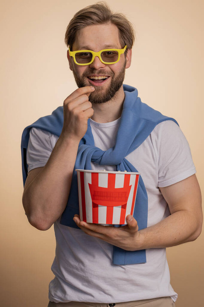 Захоплений білий чоловік їсть попкорн, дивлячись цікавий телевізійний серіал, спортивну гру, фільм, онлайн-контент соціальних медіа. Молодий хлопець насолоджується домашніми розвагами ізольований на бежевому студійному фоні
 - Фото, зображення
