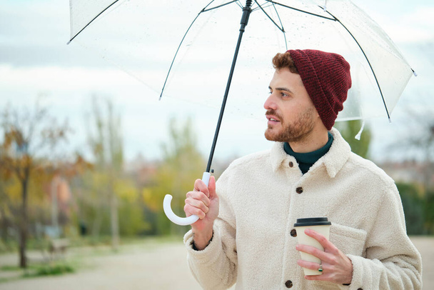 Jonge man met een koffie en transparante paraplu in de straat in de winter. - Foto, afbeelding