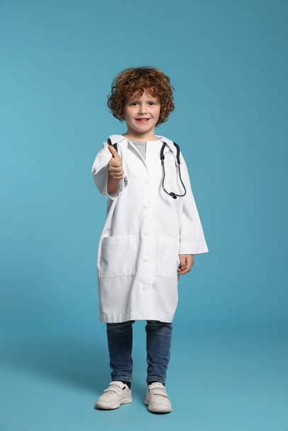 Little boy in medical uniform showing thumbs up on light blue background - Fotoğraf, Görsel