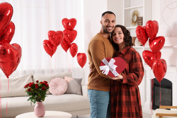 Happy couple celebrating Valentine's day. Beloved woman with gift box in room decorated with heart shaped air balloons - Foto, Bild