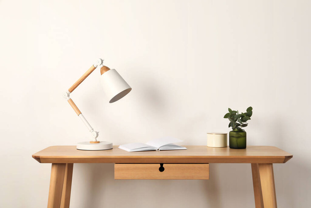 Stylish modern desk lamp, open book and plant on wooden table near white wall - Fotoğraf, Görsel