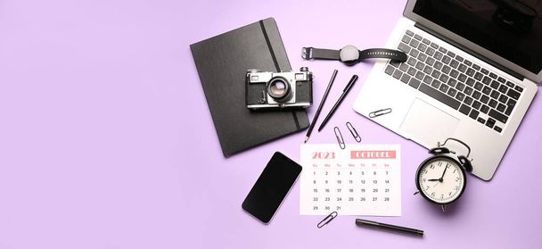 Kalender für Oktober 2023 mit Laptop, Handy, Wecker, Büropapier und Fotokamera auf fliederfarbenem Hintergrund mit Platz für Text - Foto, Bild