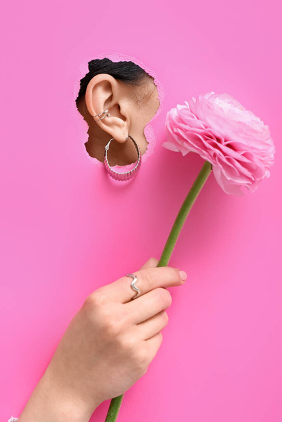 Αυτί από όμορφη νεαρή γυναίκα με κομψά σκουλαρίκια ορατά μέσα από σκισμένο ροζ χαρτί - Φωτογραφία, εικόνα