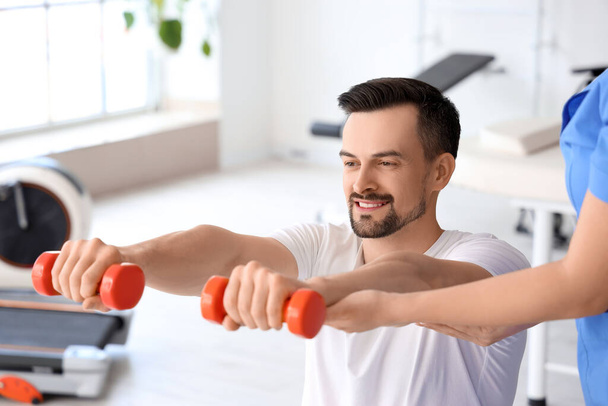 Young man training with dumbbells and physiotherapist in rehabilitation center - Фото, изображение