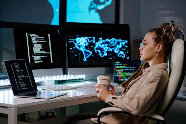 Fiatal komoly üzletasszony egy csésze kávéval ül a karosszékben a munkahelyén, és nézi a számítógép képernyőjén kódolt adatokat - Fotó, kép