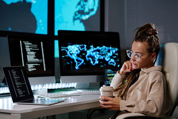 Komoly fiatal, szemüveges programozó, aki kódolt adatokat néz a laptop képernyőjén, és kávézik, miközben a munkahelyén ül a sötét irodában. - Fotó, kép