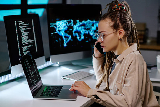 Junge fleißige IT-Ingenieur in Brille nimmt Anruf des Kunden und Überprüfung der Informationen in Laptop, während Sie durch Daten auf dem Bildschirm - Foto, Bild