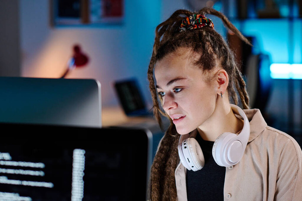 Jeune femme d'affaires avec dreadlocks assis devant l'ordinateur et regardant l'écran tout en décodant les données tard dans la soirée dans le bureau sombre - Photo, image