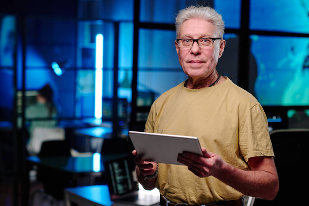Programmeur masculin senior avec tablette debout devant la caméra dans un bureau sombre et vous regardant tout en développant un nouveau logiciel - Photo, image