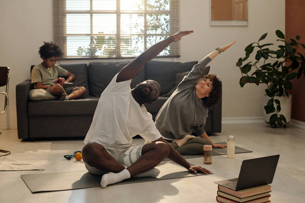 Jong intercultureel paar in activewear doen stretching oefening terwijl zitten op matten in de voorkant van de laptop en herhalen na online instructeur - Foto, afbeelding