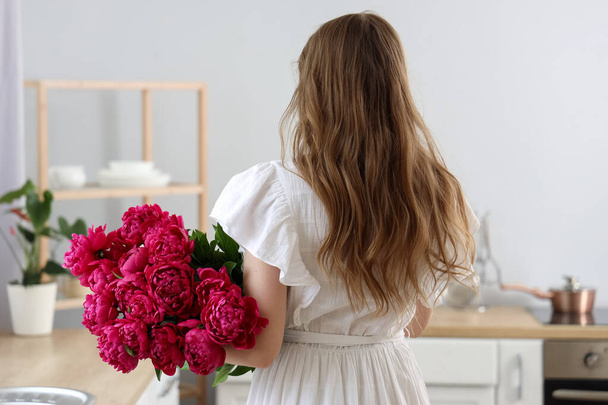 Jonge vrouw met boeket pioenroos bloemen in keuken, achteraanzicht - Foto, afbeelding