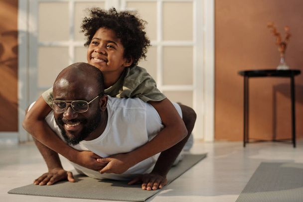 Милый веселый афроамериканец, обнимающий отца, отжимаясь на коврике во время утренней тренировки на полу в гостиной. - Фото, изображение