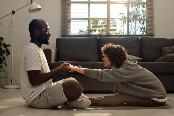 Jeune homme heureux aidant sa femme pendant l'exercice d'étirement tout en étant assis devant elle sur le tapis dans le salon et en la tenant par les mains - Photo, image