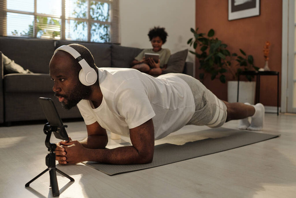 Junger Mann in Kopfhörer und Aktivkleidung wiederholt Planke nach Fitnesstrainerin auf Smartphone-Bildschirm, während er zu Hause auf Matte trainiert - Foto, Bild