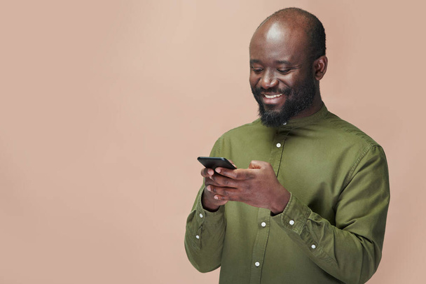 Afroamerykanin ogląda wideo na smartfonie i uśmiecha się stojąc na brązowym tle - Zdjęcie, obraz