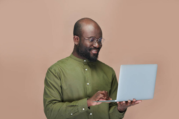 Χαμογελώντας Αφροαμερικανός κρατώντας φορητό υπολογιστή και πληκτρολογώντας στο πληκτρολόγιο, ενώ στέκεται σε καφέ φόντο - Φωτογραφία, εικόνα
