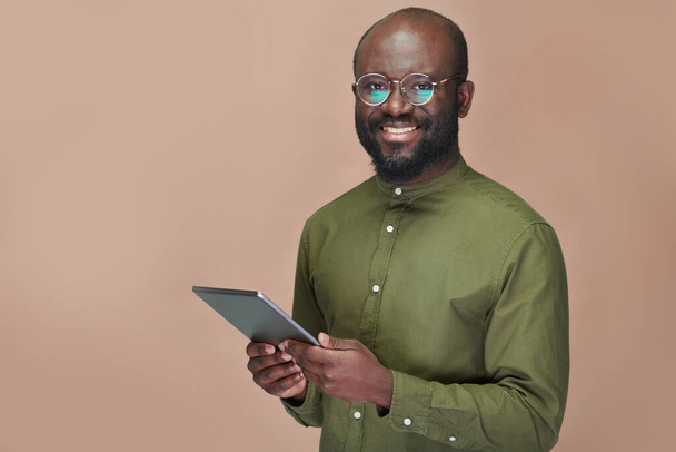 Portrait of African American man in eyeglasses smiling at camera while using tablet pc against brown background - Φωτογραφία, εικόνα