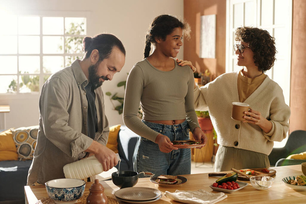 Familie van drie met geadopteerde dochter die met elkaar praten en samen ontbijten in de keuken thuis - Foto, afbeelding