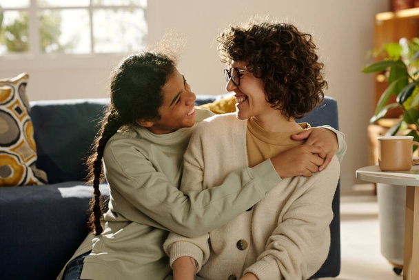 Αφροαμερικανή ευτυχισμένη έφηβη που αγκαλιάζει τη θετή της μητέρα ενώ περνούν χρόνο μαζί στο σπίτι. - Φωτογραφία, εικόνα