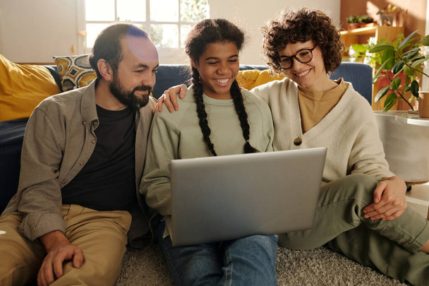 Щаслива сім'я дивиться смішне відео на ноутбуці разом зі своєю прийомною донькою під час дозвілля вдома
 - Фото, зображення