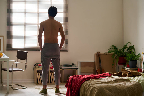 Rear view of young shirtless man worried about his problems standing alone in his bedroom - 写真・画像