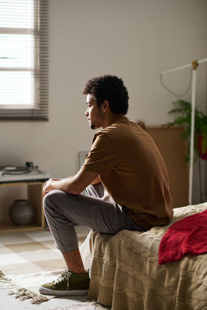 Rückansicht eines jungen Mannes, der in Gedanken auf dem Bett sitzt, während er allein in seinem Schlafzimmer bleibt - Foto, Bild
