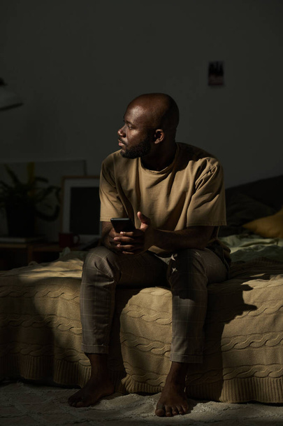 Afrikai amerikai férfi ül az ágyon mobiltelefonnal és gondolkodik valamin. - Fotó, kép