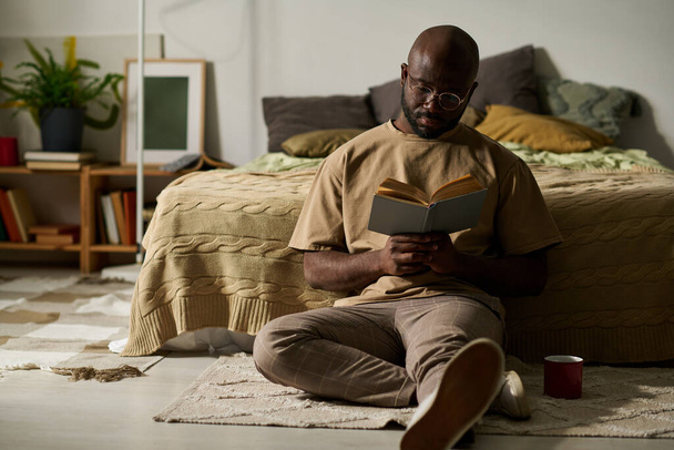 Hombre afroamericano sentado en el suelo con libro y disfrutando de la lectura en su tiempo libre en casa - Foto, Imagen