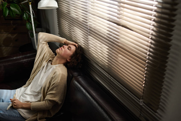 Jovem mulher morena estressada com os olhos fechados sentados em confortável sofá de couro preto pela janela e tocando a cabeça durante a sessão - Foto, Imagem