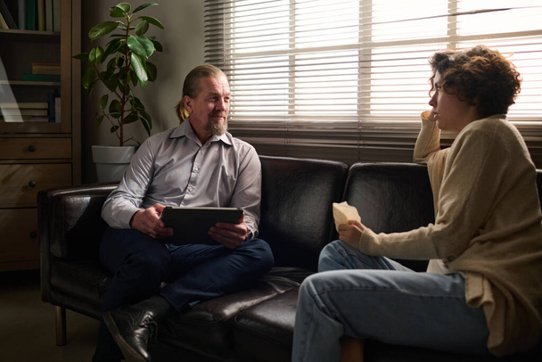 Mature confident psychotherapist with tablet communicating with young stressed female patient sitting on couch in front of him during session - Photo, Image