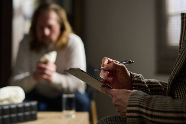 Focus op de handen van volwassen vrouwelijke psycholoog het maken van notities in document terwijl zitten in de voorkant van de camera tegen depressieve mannelijke patiënt - Foto, afbeelding