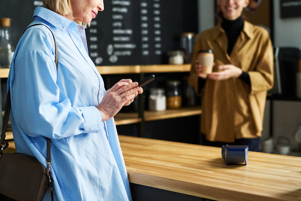 Mavi gömlekli olgun bir kadın kafeteryada tezgah başında beklerken akıllı telefon kullanıyor ve katibin kahve hazırlamasını bekliyor. - Fotoğraf, Görsel