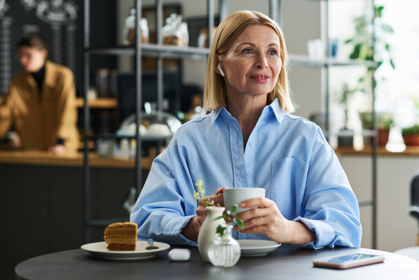 Szczęśliwa blondynka dojrzała kobieta ze słuchawkami słuchającymi muzyki, siedząca przy stole w kawiarni i pijąca herbatę lub kawę z ciastem - Zdjęcie, obraz