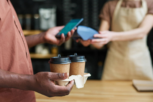 Close-up of African American mature male customer holding two cups of coffee and paying for his order by smartphone by counter desk - Fotó, kép