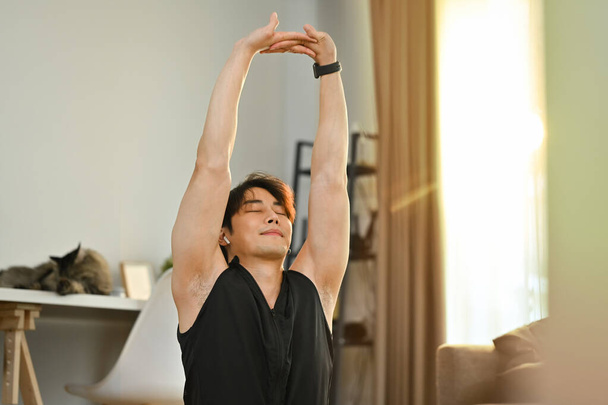 Zadowolony Azjata w sportowym stroju rozciągający ramiona odpoczywający po treningu domowym. Zdrowy styl życia i koncepcja fitness. - Zdjęcie, obraz
