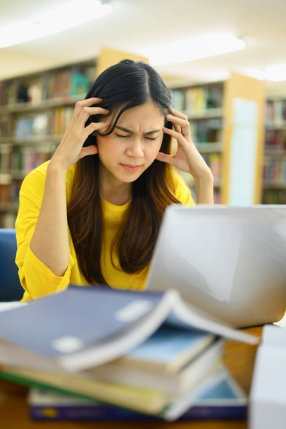 Wyczerpana studentka siedząca w bibliotece z laptopem, cierpiąca na ból głowy lub zmęczona nauką do egzaminu na studia. - Zdjęcie, obraz